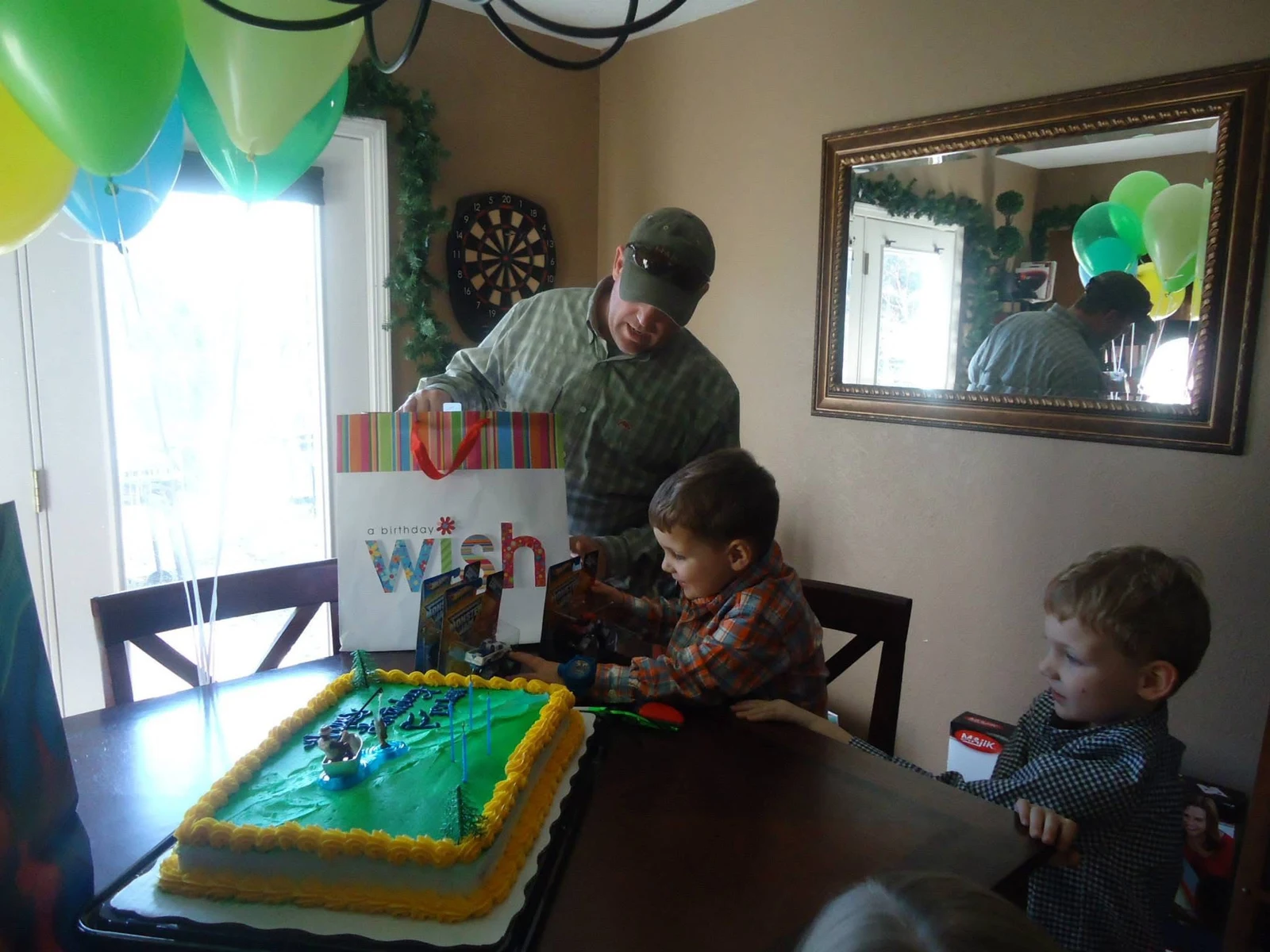 A birthday table with green balloons, a cake, and children opening gifts.