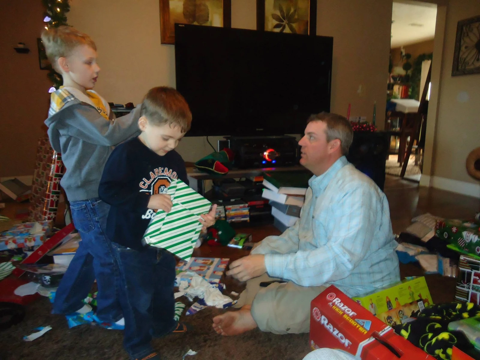 Two children opening Christmas presents beside an adult in the living room.